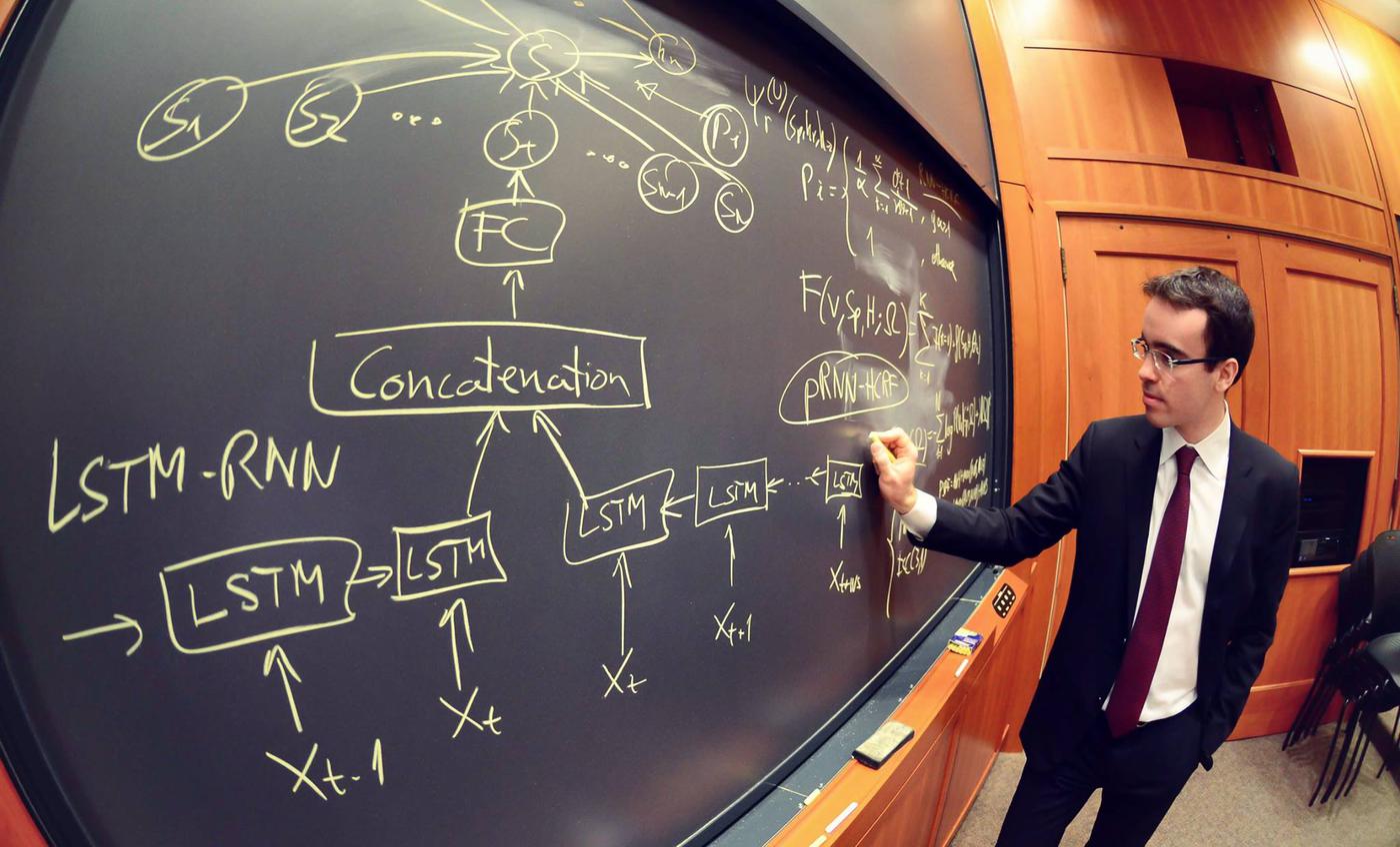 Daniel Lopez-Martinez, in a suit and tie, drawing an LSTM recurrent neural network architecture in chalk on the board of a Harvard Business School classroom..